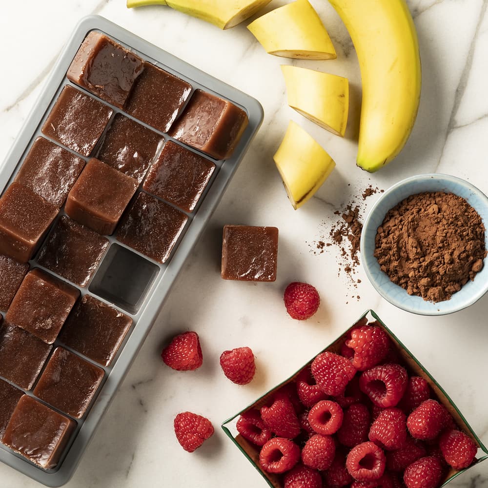 Chocolates, frozen bananas and raspberries, and dutch processed cocoa powder on a countertop to make a chocolate raspberry smoothie